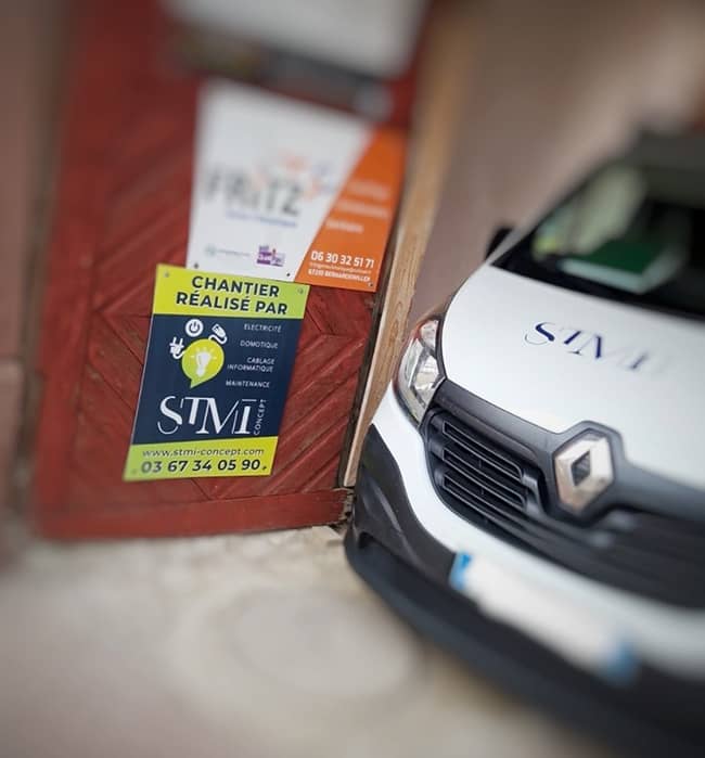 Une camionnette Renault blanche est garée près d'une porte en bois avec une signalisation affichant les coordonnées de STMI